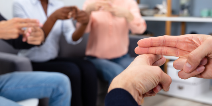 Das Bild zeigt mehrere Personen, die in einem Stuhlkreis sitzen. Alle Personen legen Mittelfinger und Zeigefinger der rechten Hand auf den Zeigefinger der linken Hand und zeigen damit eine Gebärde.