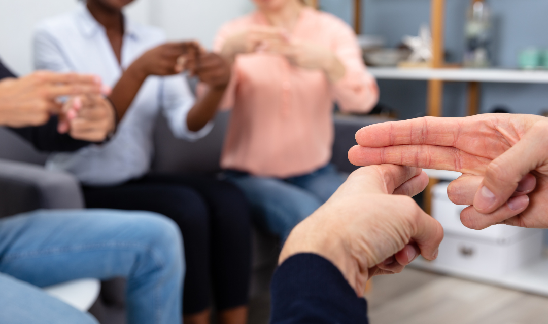 Das Bild zeigt mehrere Personen, die in einem Stuhlkreis sitzen. Alle Personen legen Mittelfinger und Zeigefinger der rechten Hand auf den Zeigefinger der linken Hand und zeigen damit eine Gebärde.